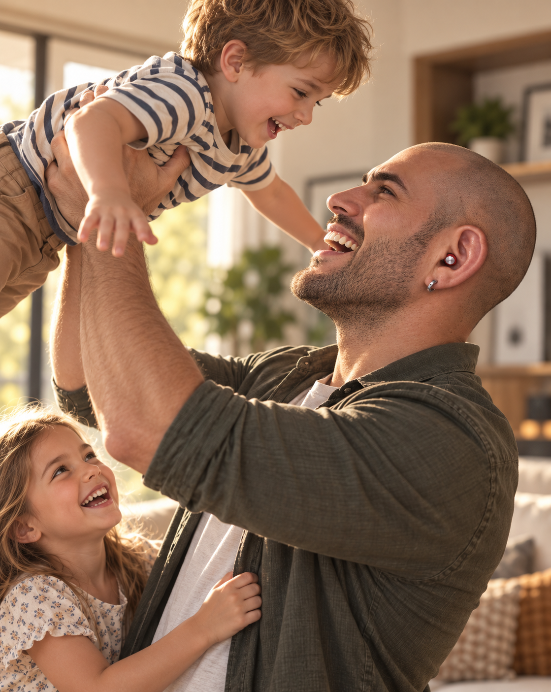 A smiling man lifts a young boy above his head in a bright living room, while a young girl looks on. The man wears a dark olive green shirt and a Roam Everyday Invisible Earplug in Crimson is visible in his ear.