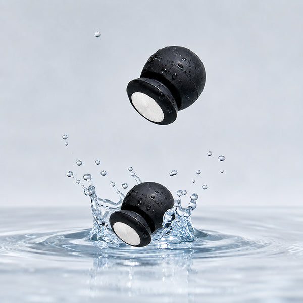 Two black silicone earbud covers with white inner discs floating in water, with one rising and one splashing down, against a light gray background.